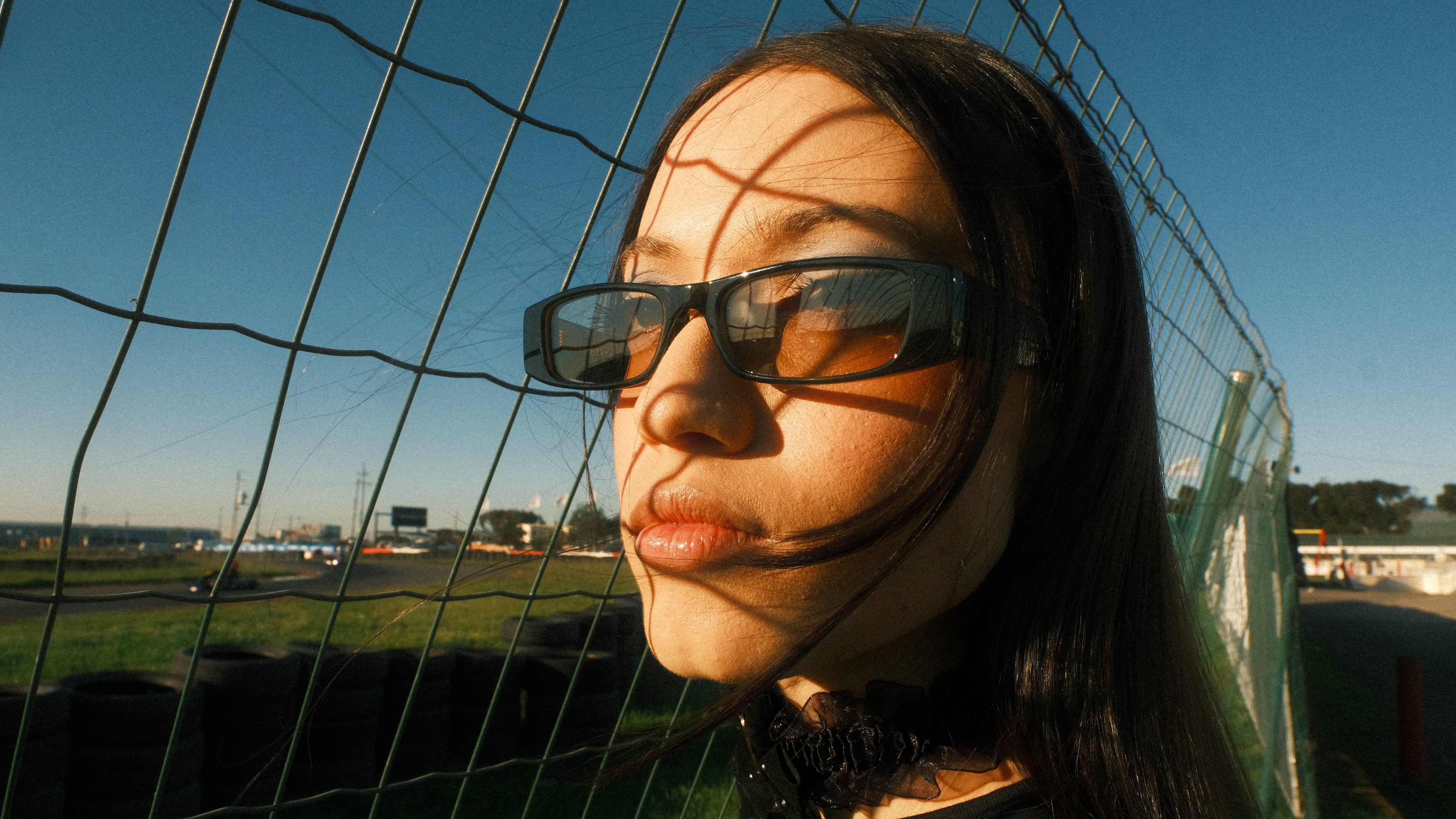 A close-up portrait of a woman wearing dark rectangular sunglasses, standing outdoors behind a wire fence with strong sunlight casting shadows across her face. She has long dark hair and is wearing a black top.